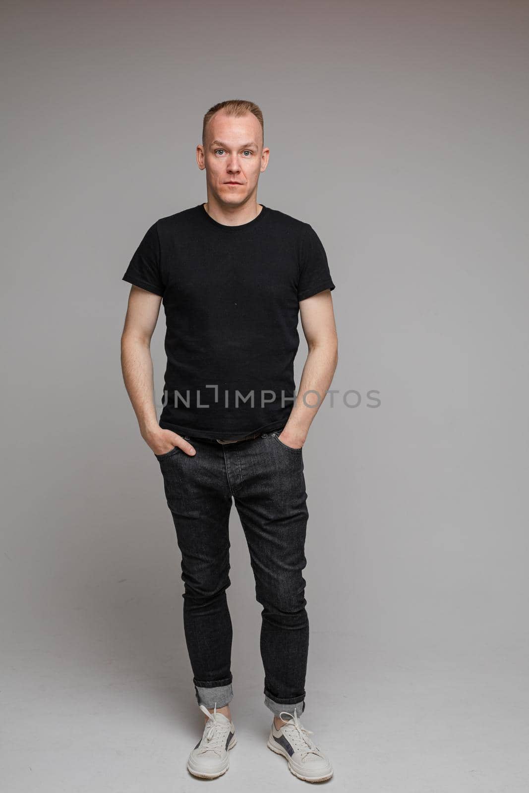 picture of attractive man wearing a black t-shirt and jeans stands with hands in pockets isolated on grey background