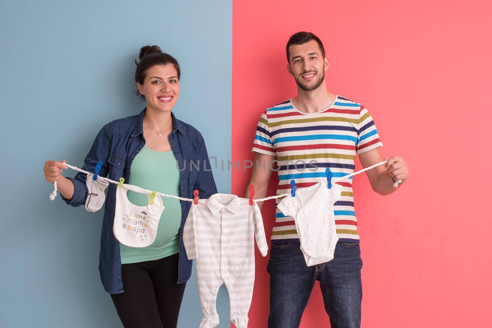 Beautiful pregnant woman and her husband expecting baby holding baby bodysuits and smiling over colorful background