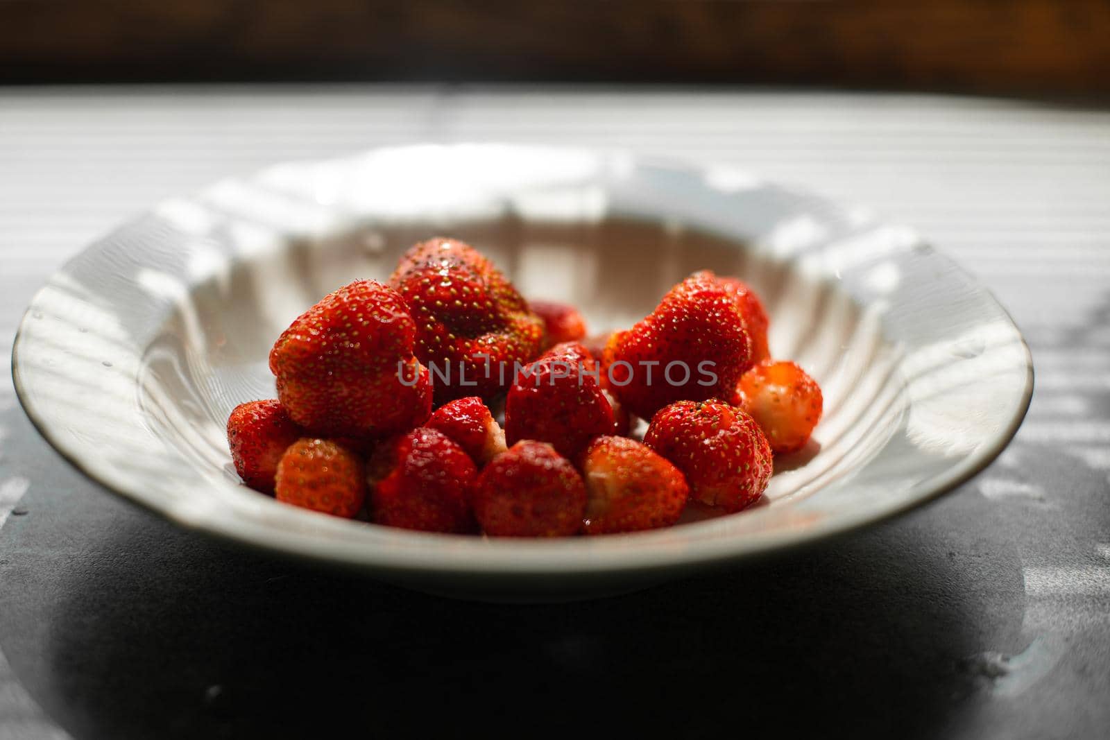 Many juicy fresh ripe red strawberry berries lie in a white ceramic plate on the table under bright sunlight by StudioLucky