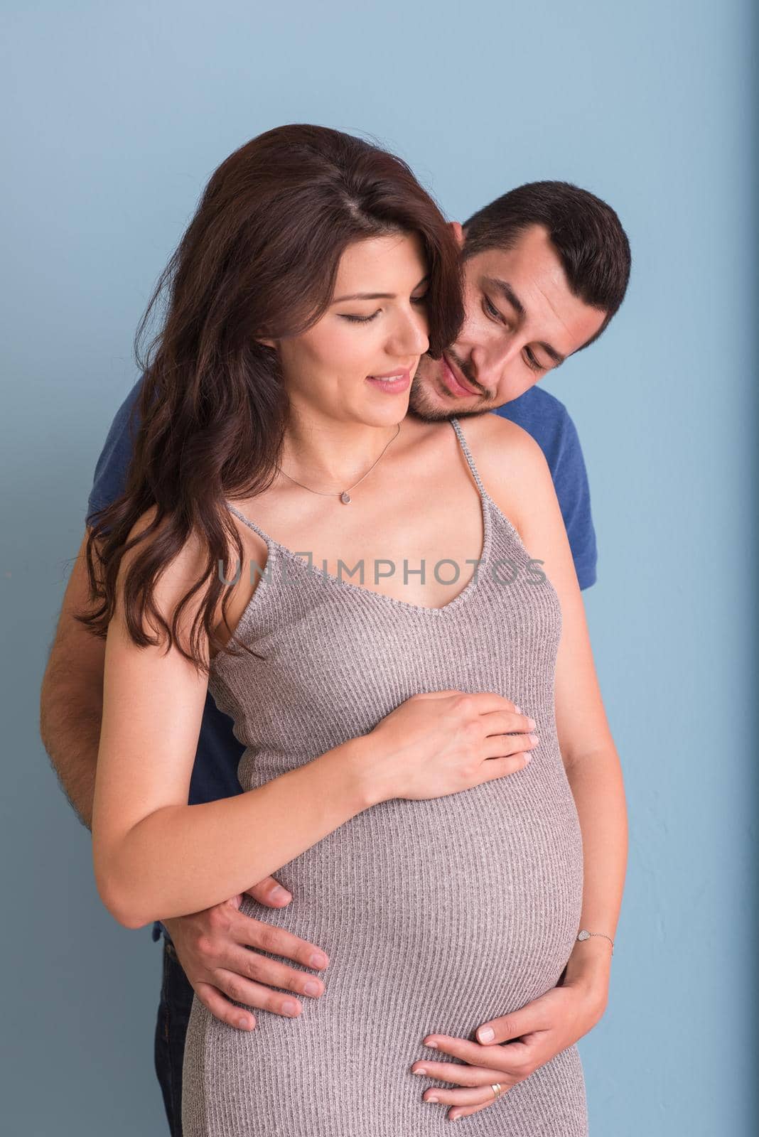 Portrait of a happy young couple,man holding his pregnant wife belly isolated over blue background