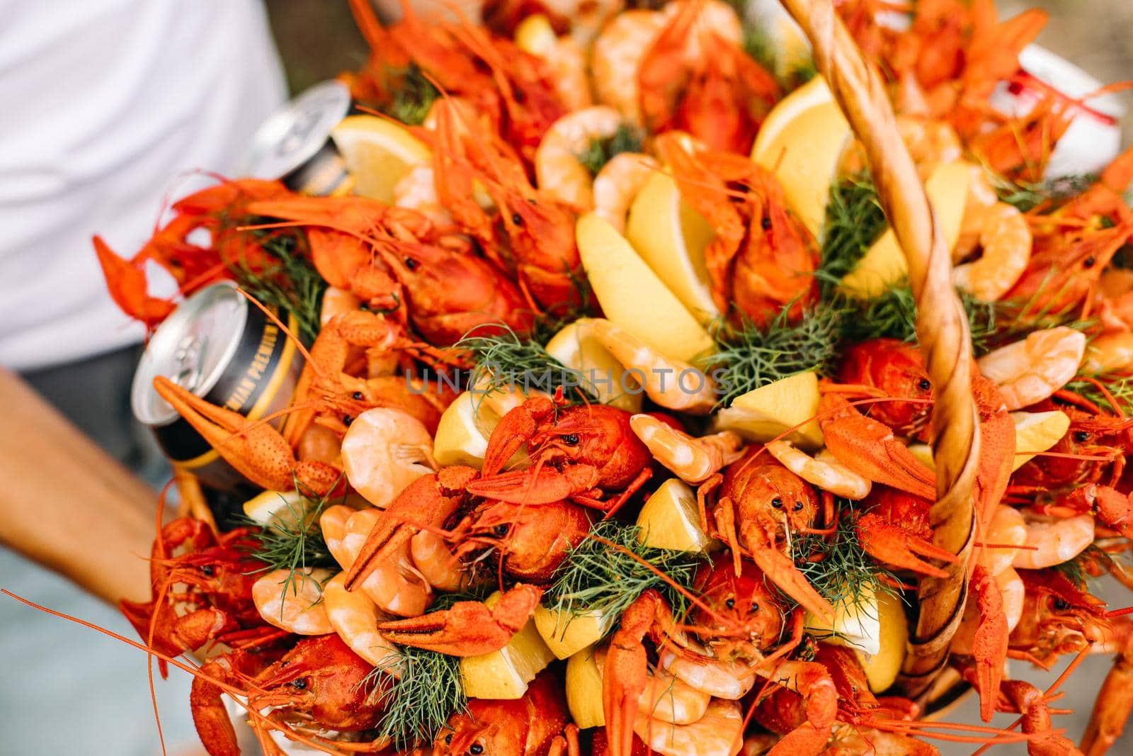 Close-up view over a festive basket full of fresh and tasty crayfish, shrimps and beer cans decorated with sliced lemon and bunches of dill. Unrecognizable person with a basket full of seafood and beer.