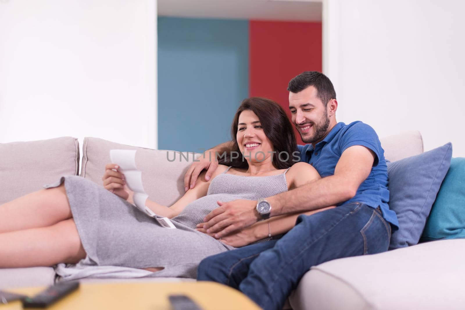 Young pregnant couple looking baby's ultrasound photo while relaxing on sofa at home