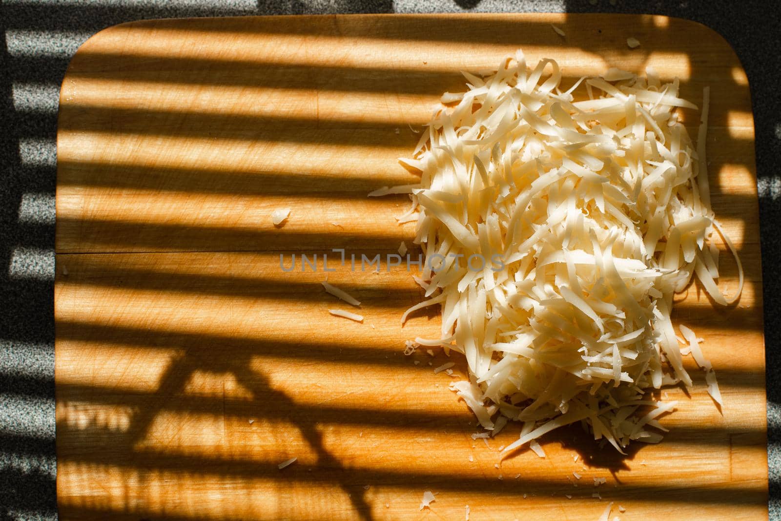 Stock photo of pile of fresh yellow grated cheese on wooden cutting board on kitchen table covered with blinds shade. Stripes from blinds on the window casting over board with shredded cheese.