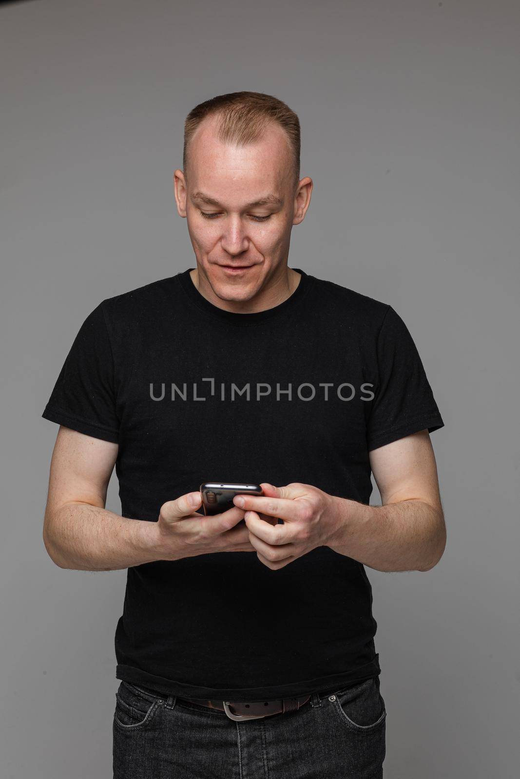 Studio portrait of cheerful blond Caucasian man in black t-shirt and jeans holding hands in pockets and smiling at camera on grey background.