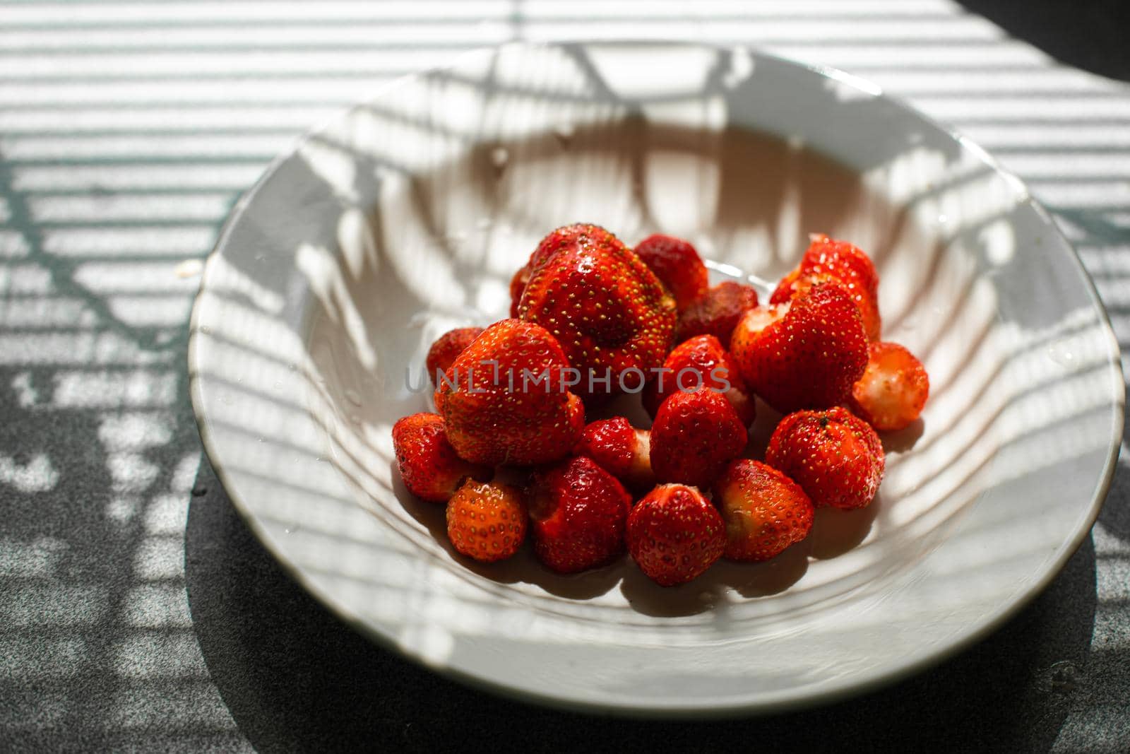 picture of juicy fresh ripe red strawberries in a white ceramic plate on the table under bright sunlight in a village