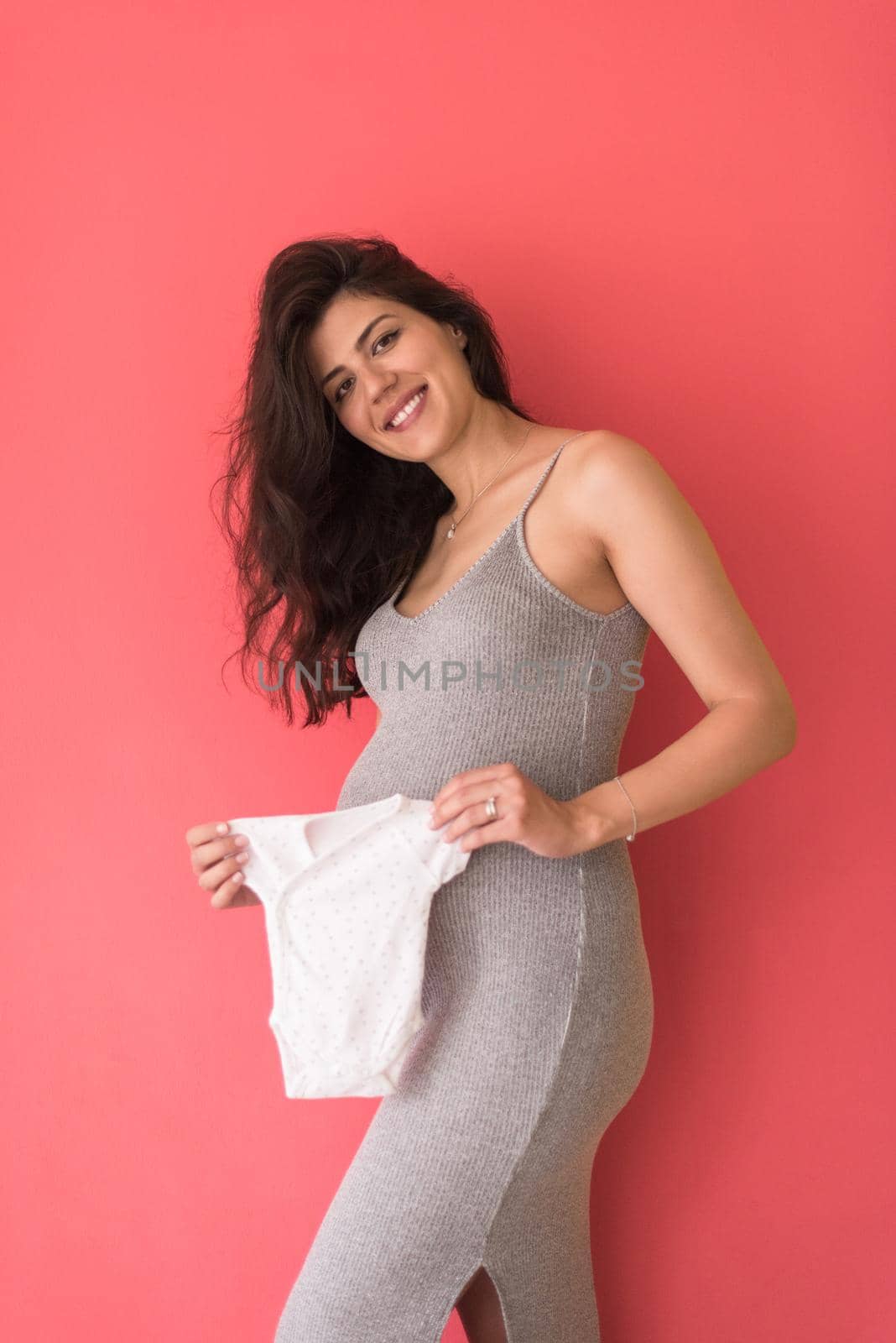 happy pregnant woman holding cute baby bodysuit, isolated on red background, baby expectation concept