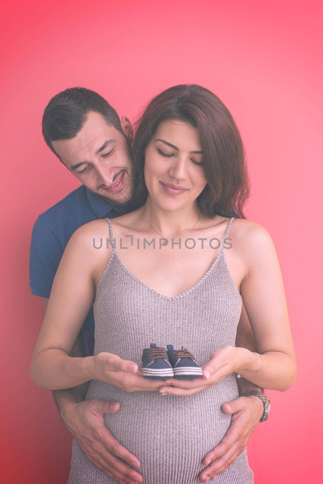 young  pregnant couple holding newborn baby shoes isolated on red background,family and parenthood concept