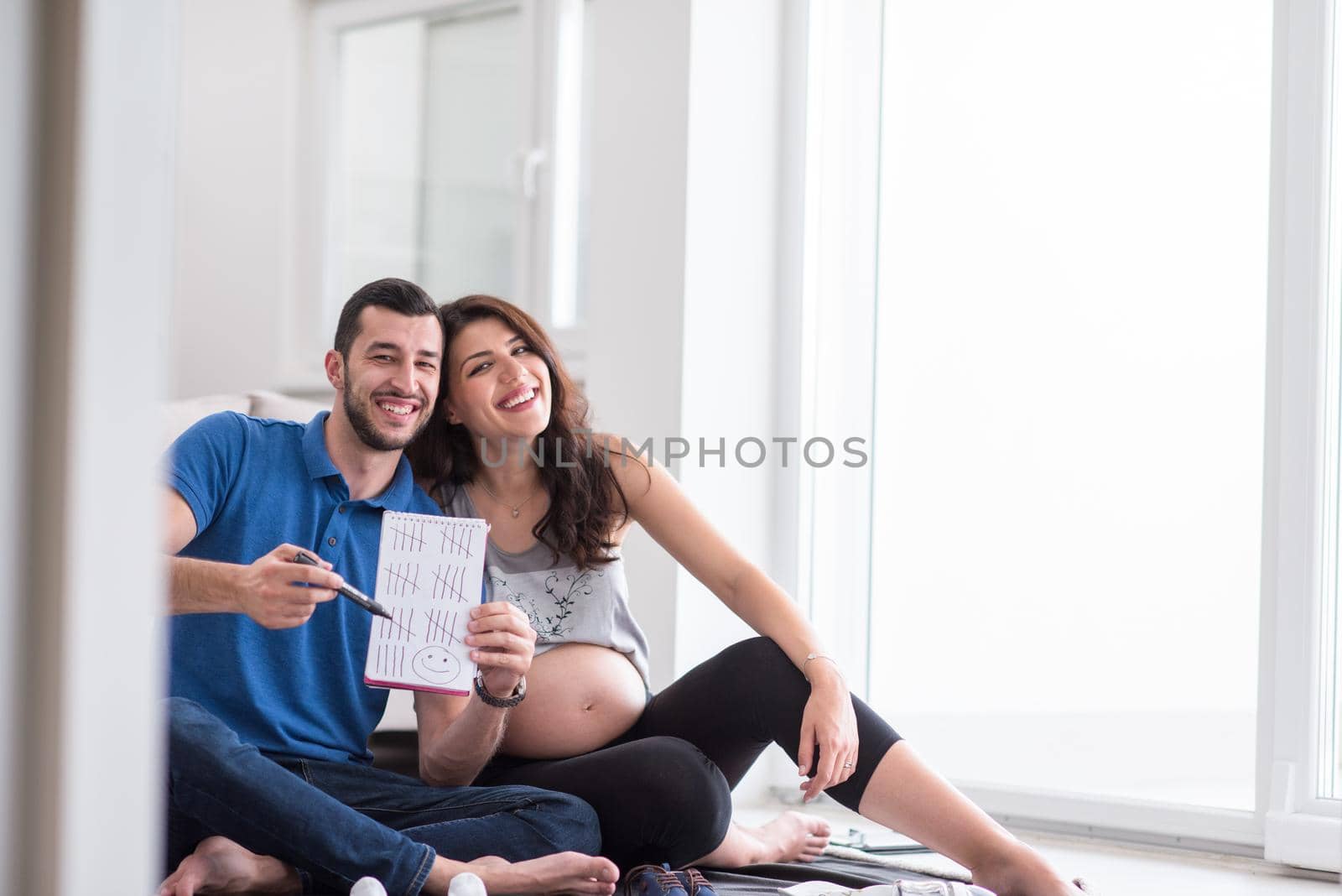 a happy pregnant couple checking a list of things for their unborn baby at home on the floor