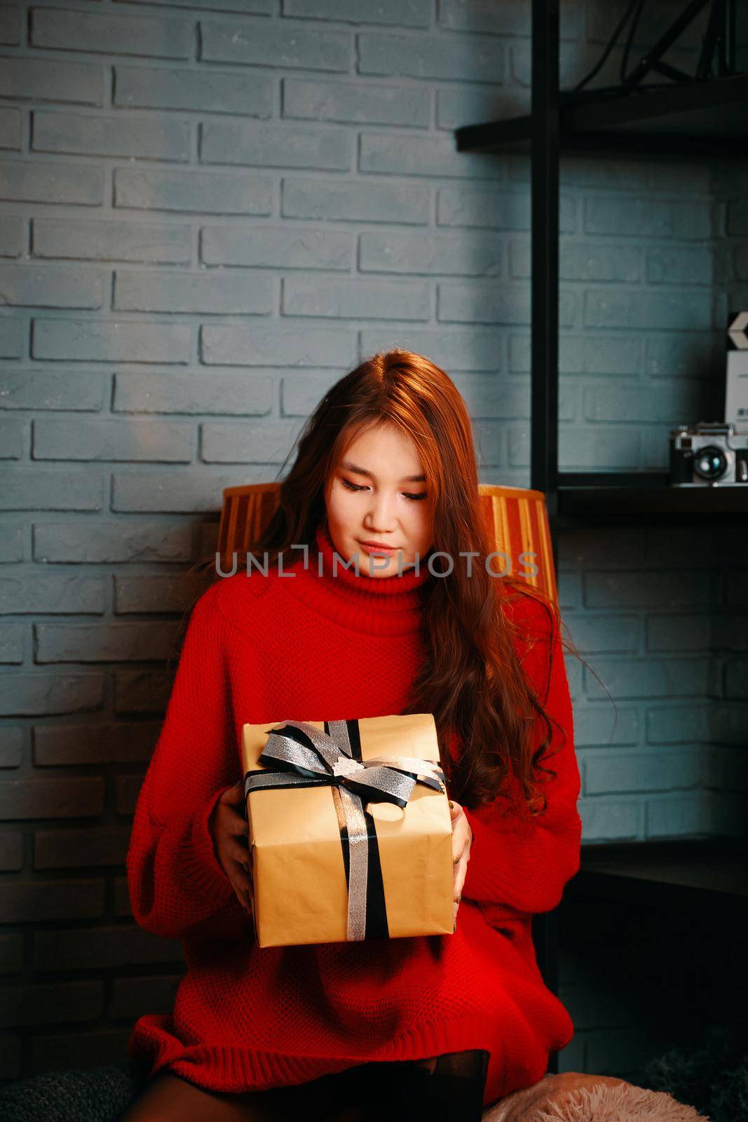 Cute woman with gift box. Asian girl in red knitted sweater. Festive mood. Gray brick wall on the background. Winter holidays for Christmas. New year mood.