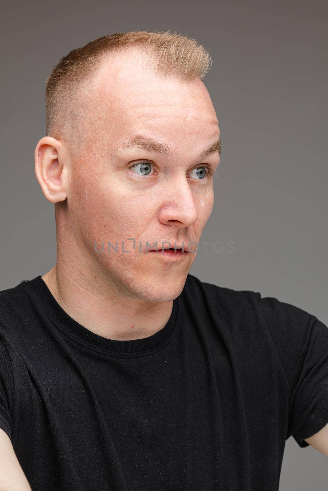 Portrait of astonished blond Caucasian man in black talking to someone showing irritation and bewilderment with cropped arms up on grey background.