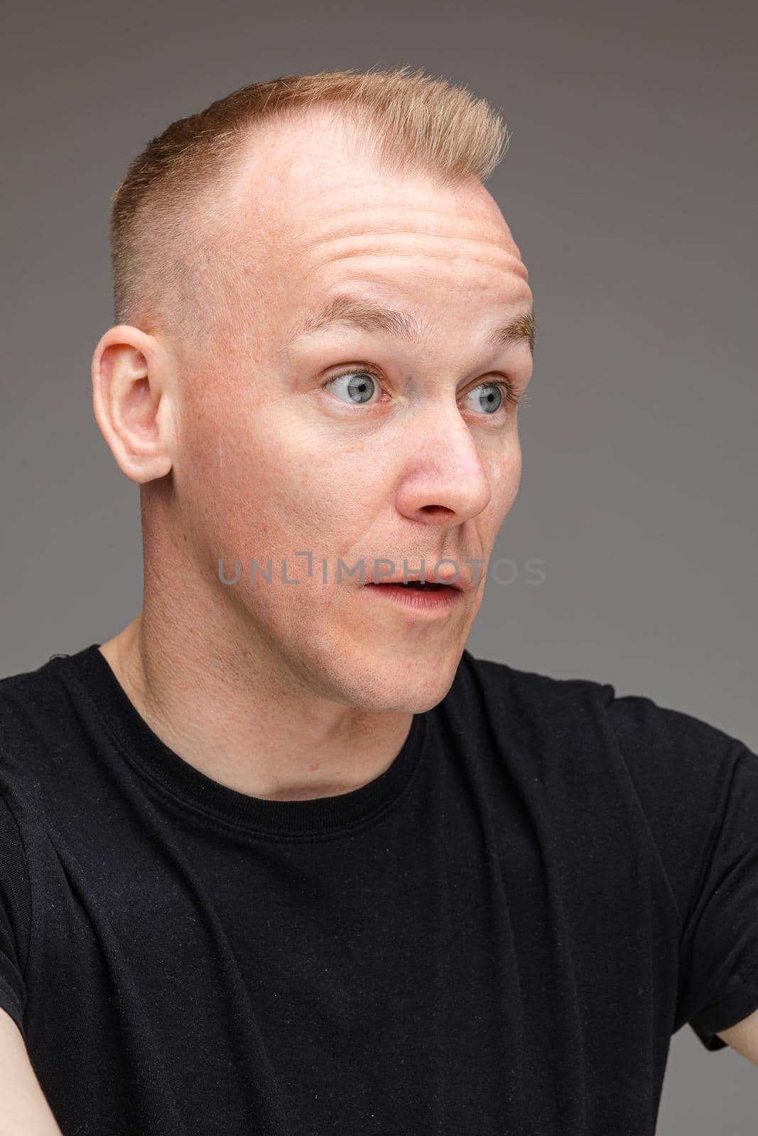 portrait of beautiful caucasian male in black t-shirt looks at something interesting with big eyes and open mouth isolated on grey background by StudioLucky