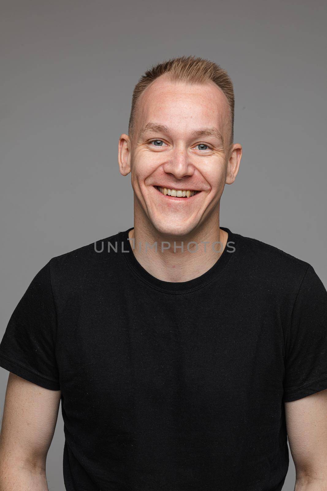 Studio portrait of adult man with short blond hair wearing casual black t-shirt smiling cheerfully at camera with friendly look. Cutout on grey background.