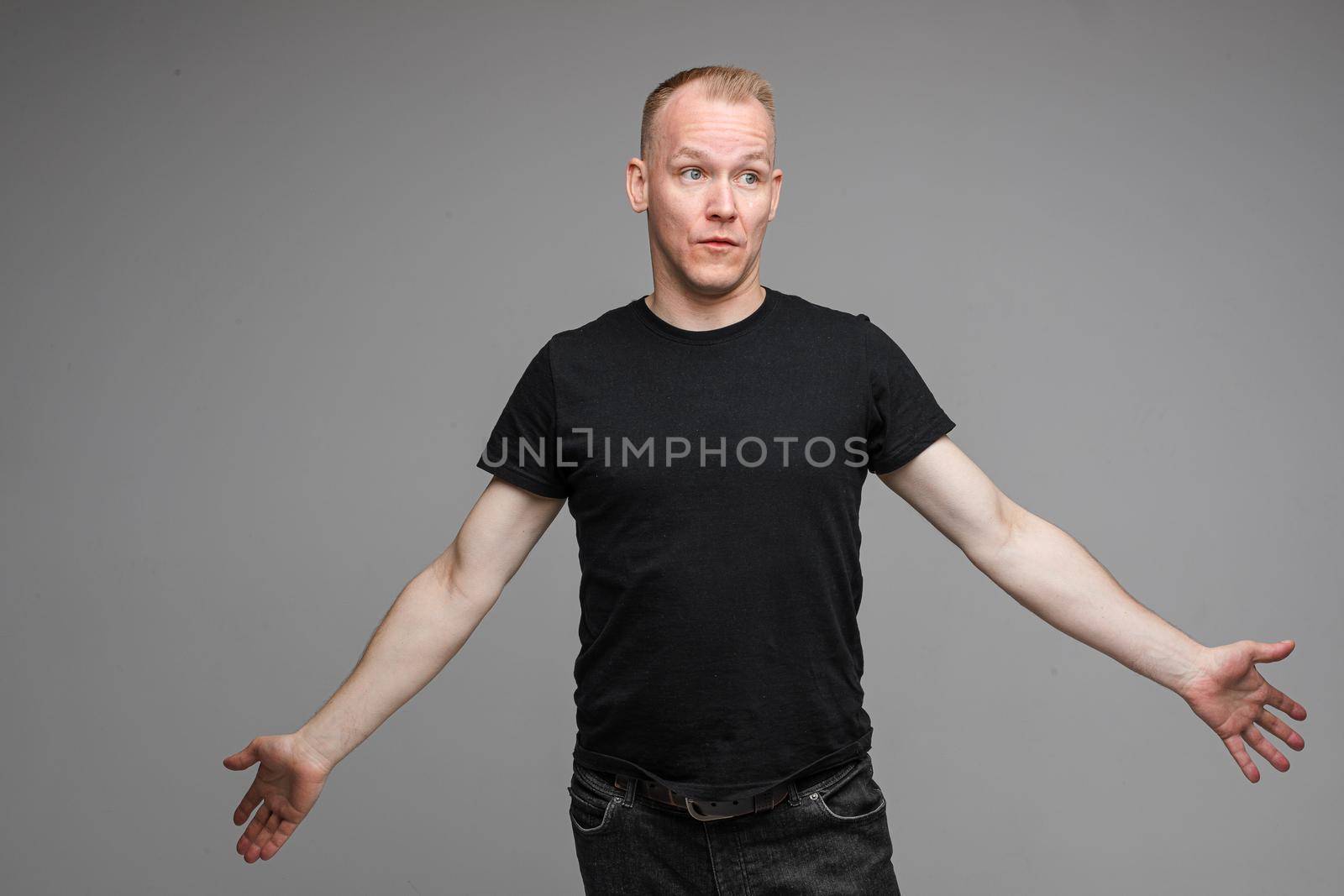 Studio portrait of handsome adult Caucasian man with short blond hair in black t-shirt and dark jeans looking away with puzzled face and outstretched arms.