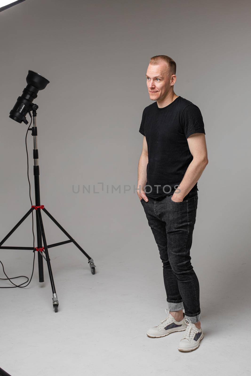 Full length portrait of stylish Caucasian man with short blond hair wearing casual black t-shirt, dark jeans and white sneakers standing with hands in pockets. Studio light in background.