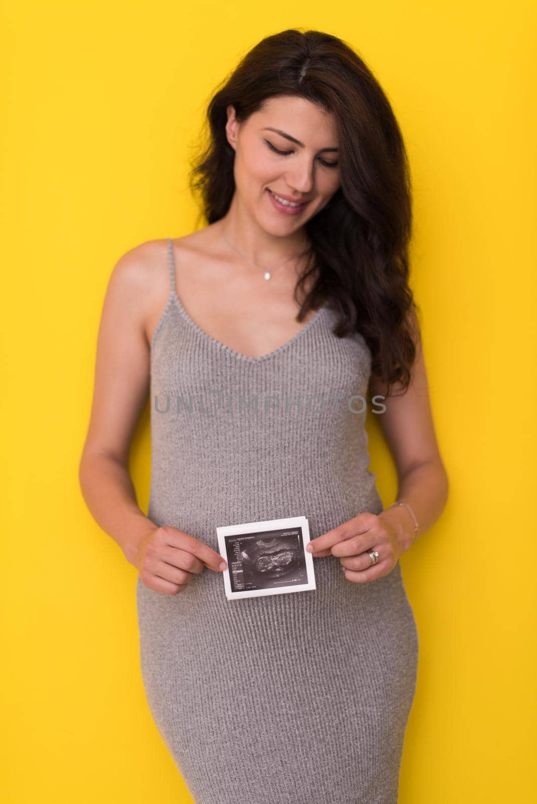 smiling pregnant woman showing ultrasound picture of her unborn baby isolated on yellow background