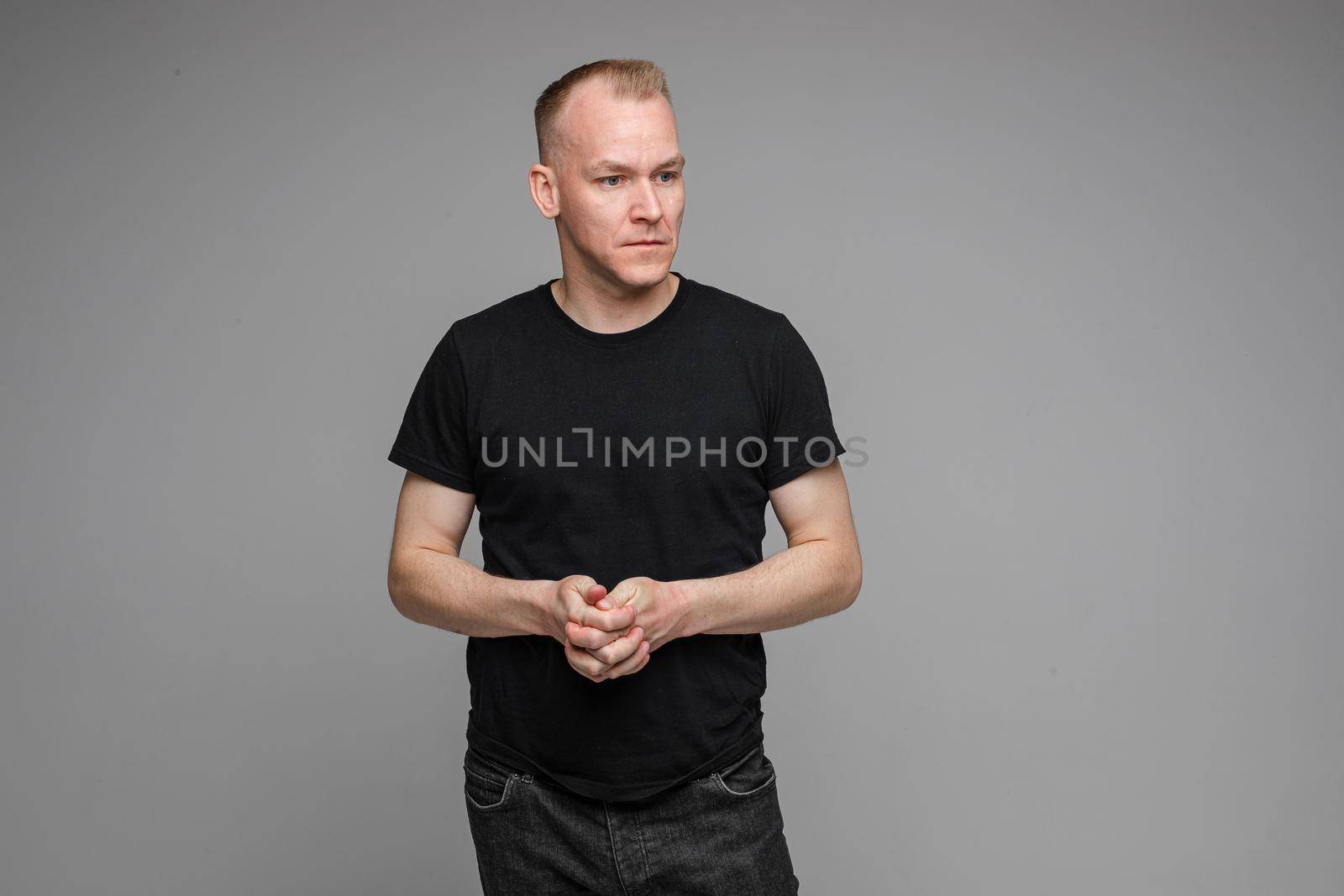 Portrait of serious blond Caucasian man in black t-shirt and dark jeans holding hands with fingers crossed looking attentively away at someone. Isolate on grey background.