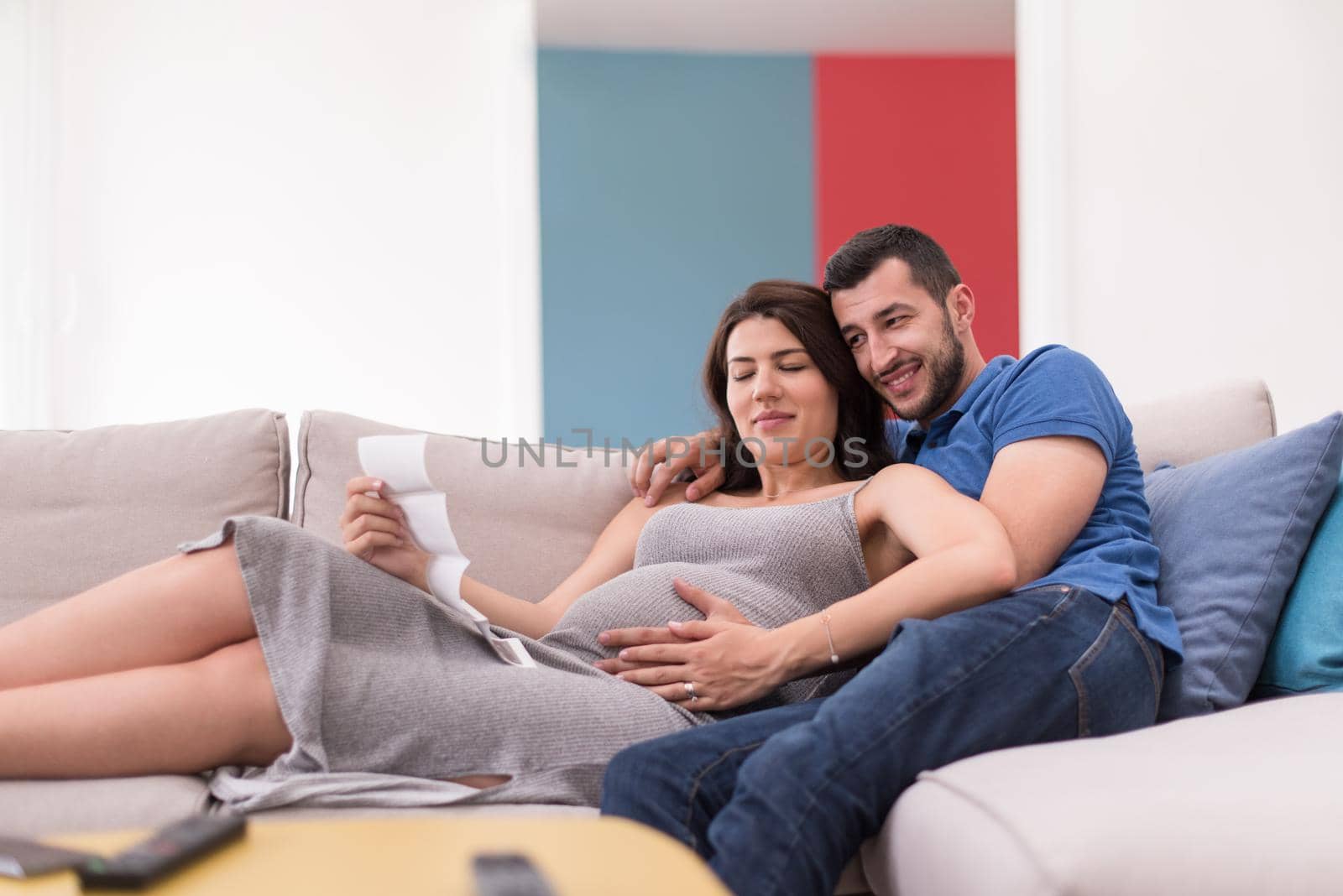 Young pregnant couple looking baby's ultrasound photo while relaxing on sofa at home