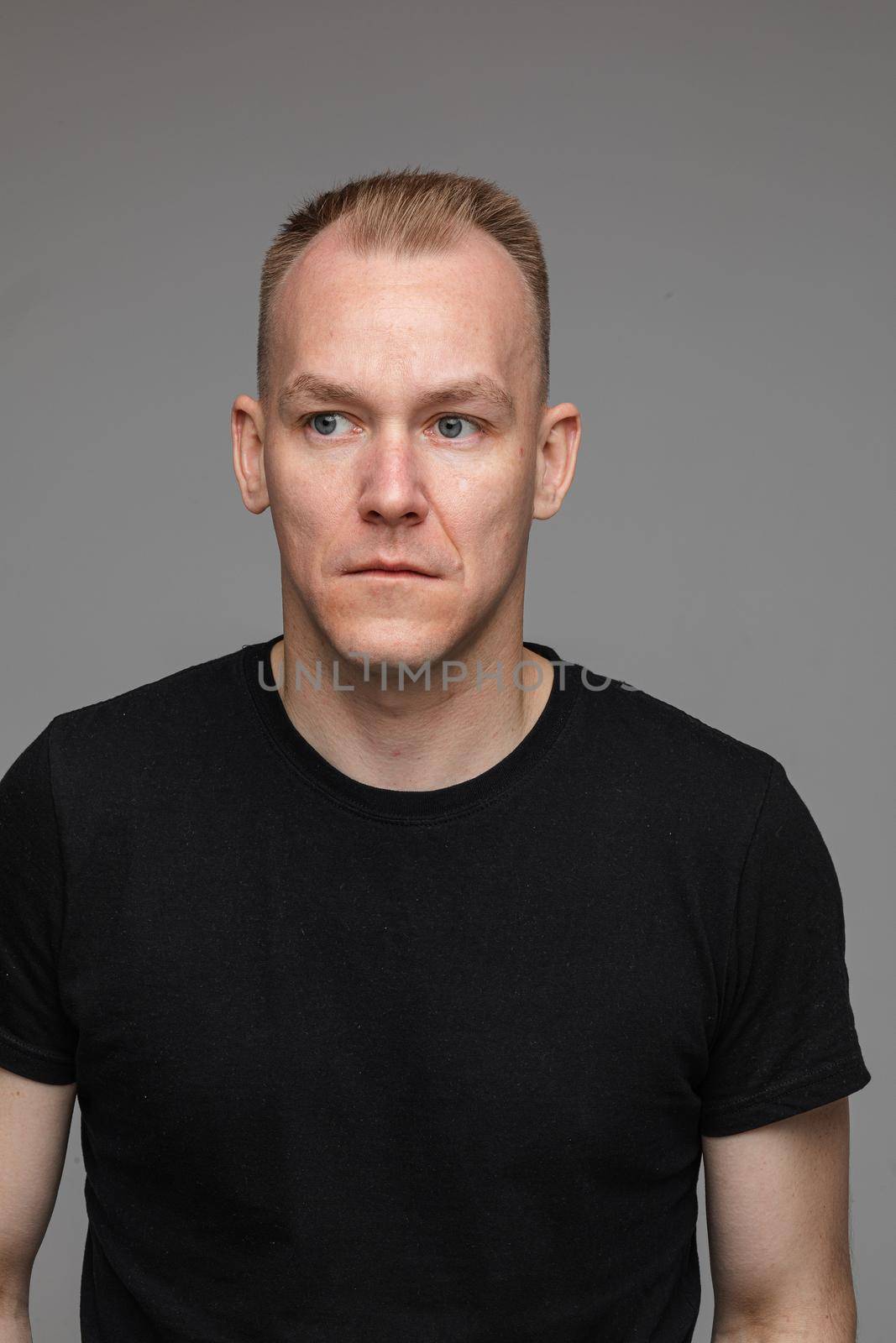 Studio portrait of cheerful blond Caucasian man in black t-shirt and jeans holding hands in pockets and smiling at camera on grey background.