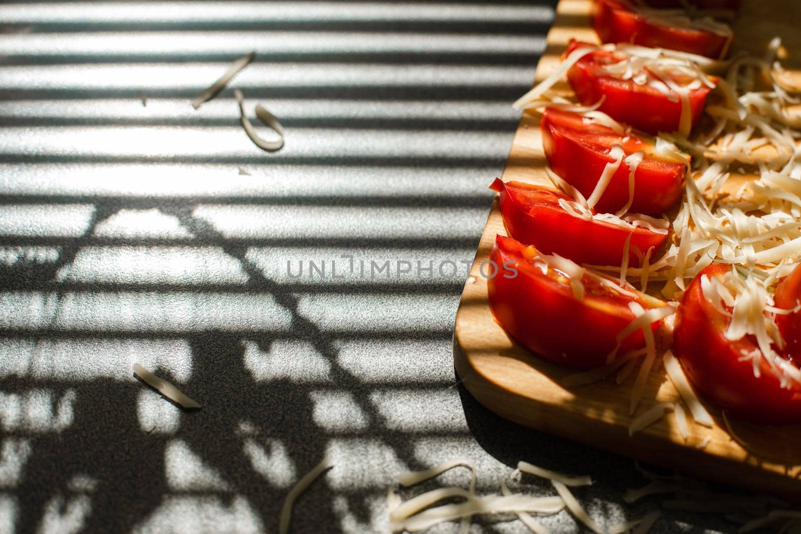 Stock photo in close up of sliced tomatoes with shredded cheese on wooden board in sunlight. Blinds shadow over the table. Cooking process.