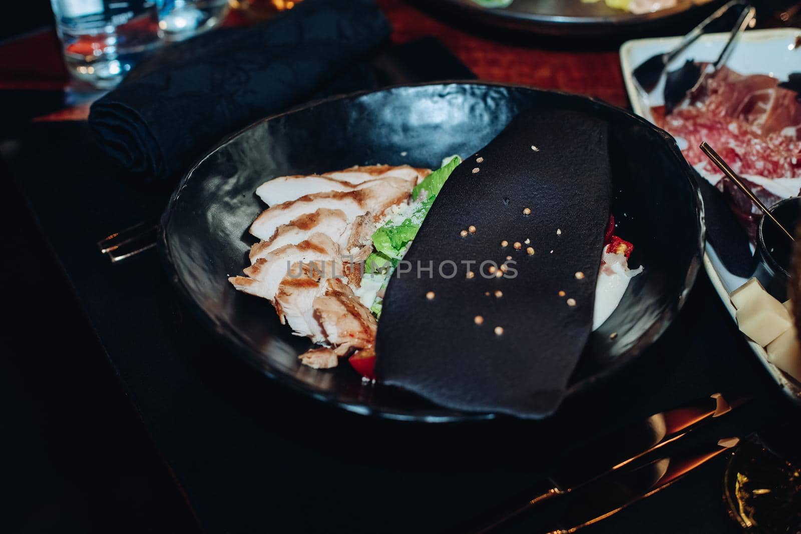 Stock photo of delicious fresh Caesar salad with chicken slices served with black slice with sesame seeds in bowl on black table.