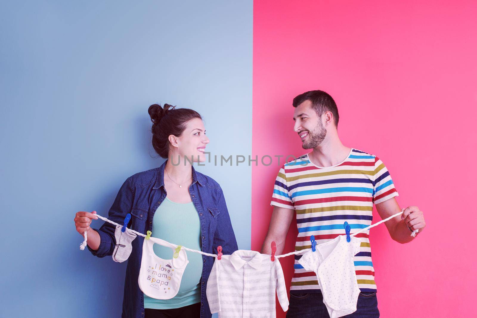 Beautiful pregnant woman and her husband expecting baby holding baby bodysuits and smiling over colorful background