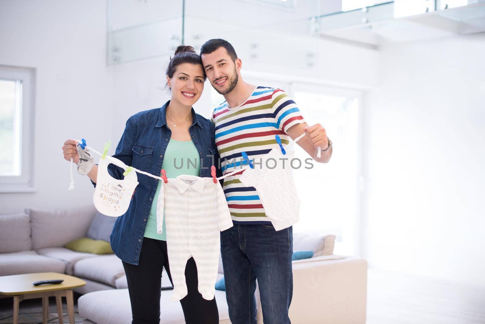 Beautiful pregnant woman and her husband expecting baby holding baby bodysuits and smiling at home