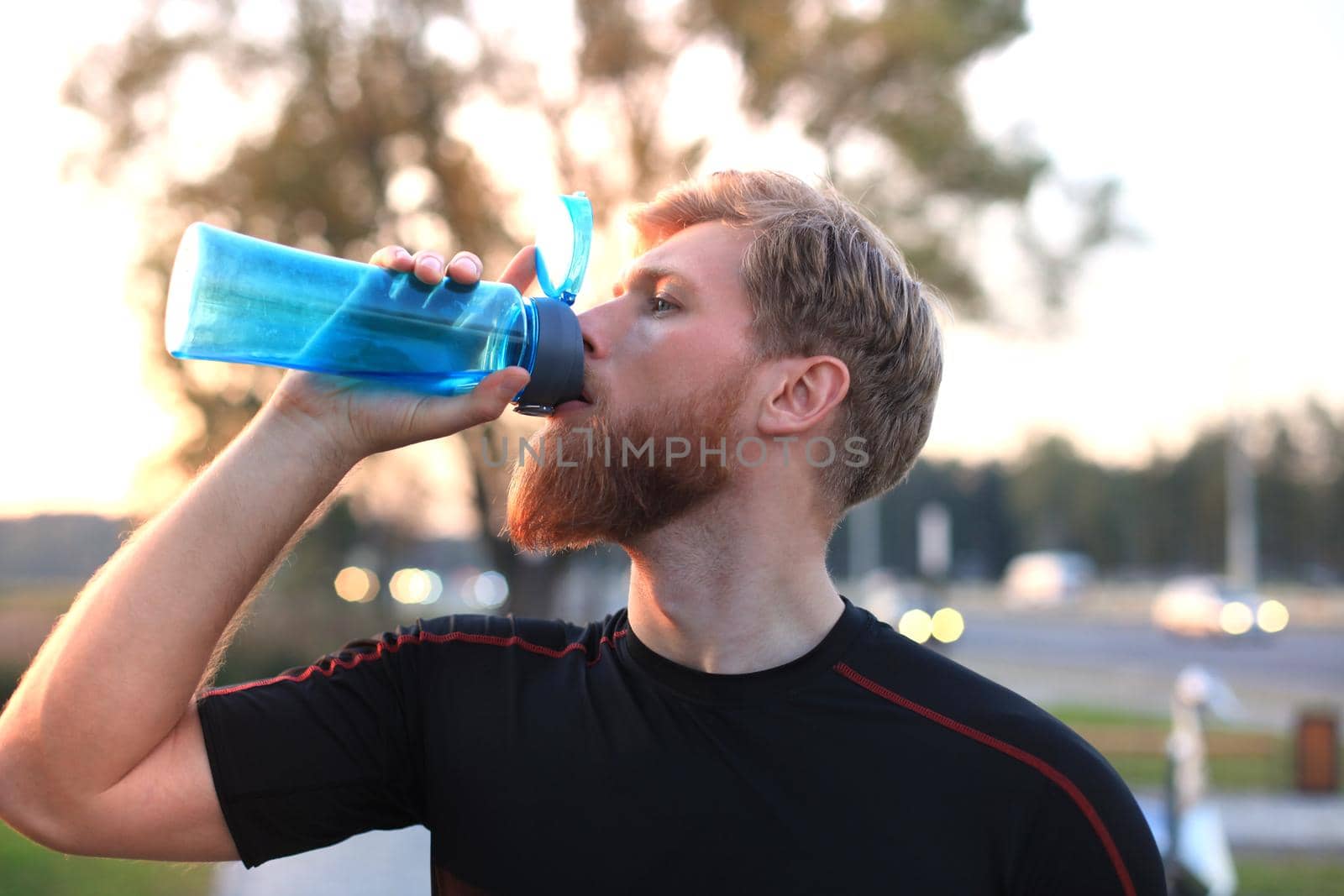 Handsome adult man drinking water from fitness bottle while standing outside, at sunset or sunrise. Runner