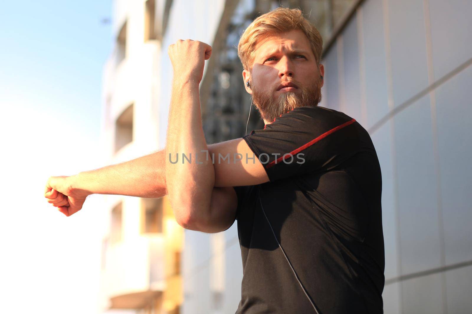 Handsome young man doing stretching exercises before running while standing outdoors by tsyhun