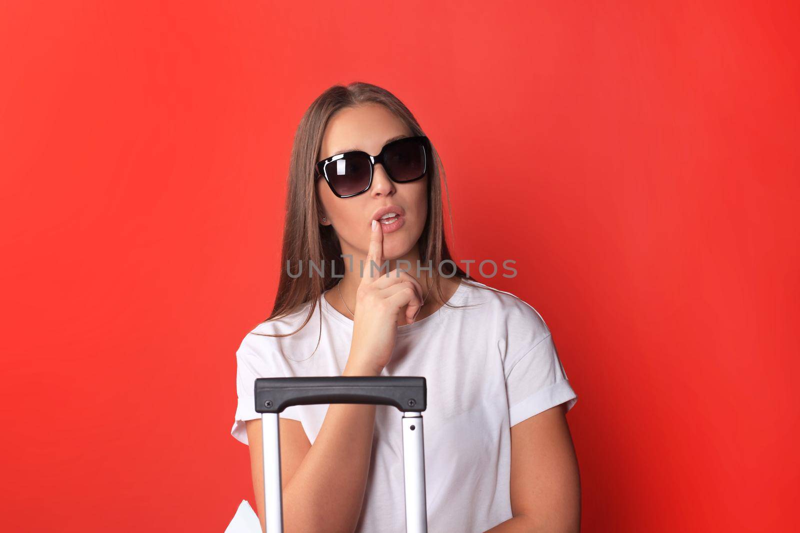 Young tourist girl in summer casual clothes, with sunglasses, red suitcase, isolated on red background.