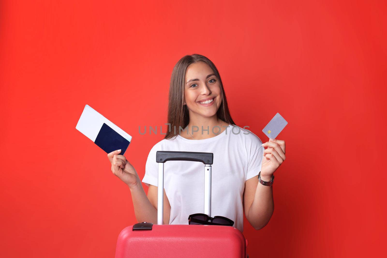 Tourist girl in summer showing plastic credit card, with sunglasses, red suitcase, passport isolated on red background. by tsyhun