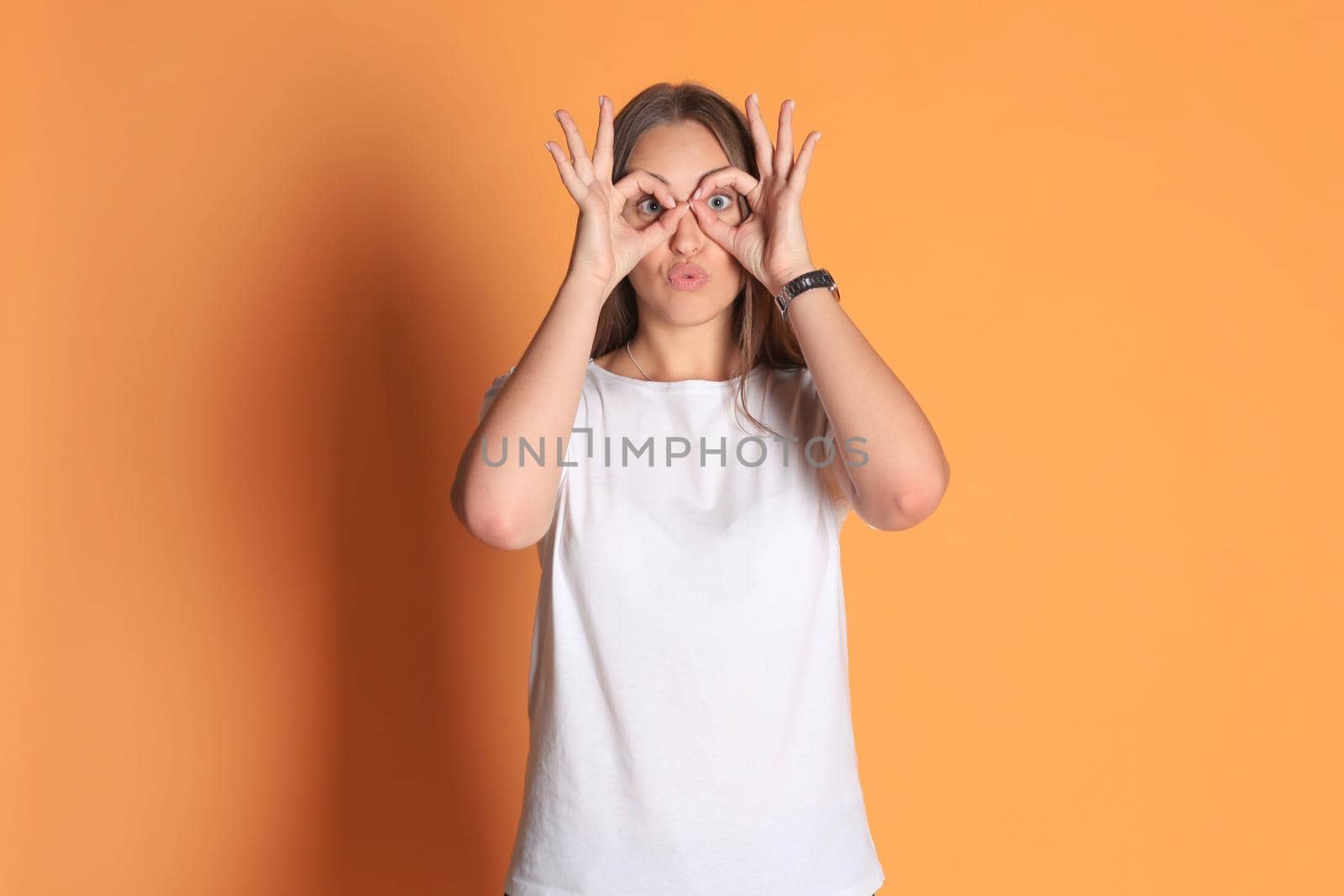 Young woman in basic clothing smiling looking at camera through holes made with fingers isolated over yellow background. by tsyhun