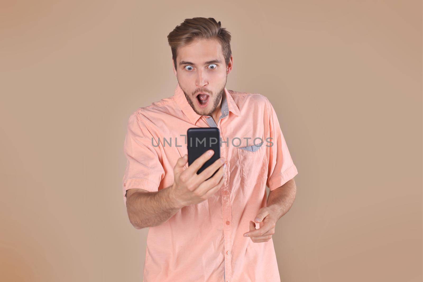 Shocked young man standing isolated over beige background, using mobile phone