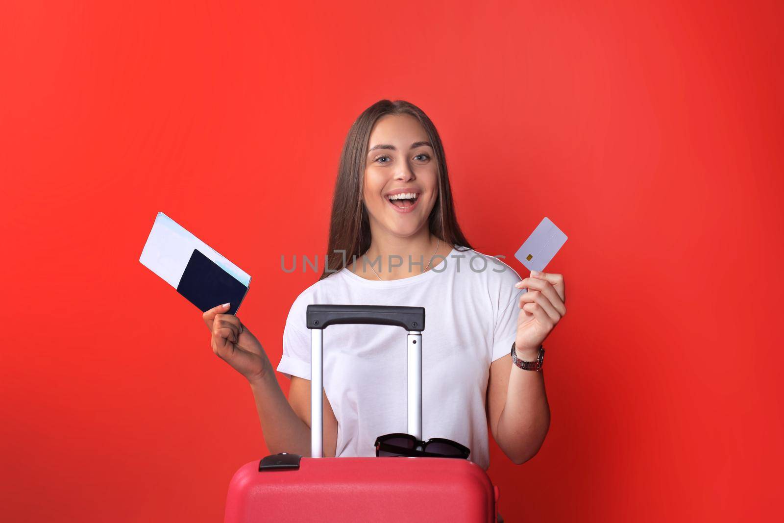 Young tourist girl in summer casual clothes, with sunglasses, red suitcase, passport isolated on red background.