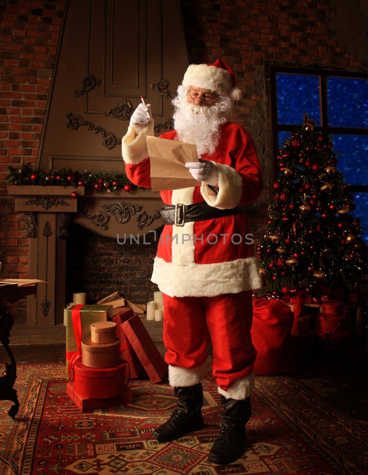 Santa Claus standing at his room at home near Christmas tree and big sack and reading Christmas letter or wish list. by tsyhun