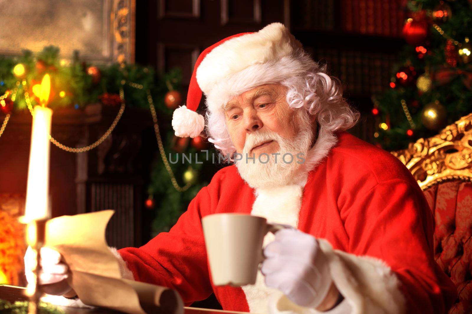 Portrait of happy Santa Claus sitting at his room at home near Christmas tree and reading Christmas letter or wish list. by tsyhun