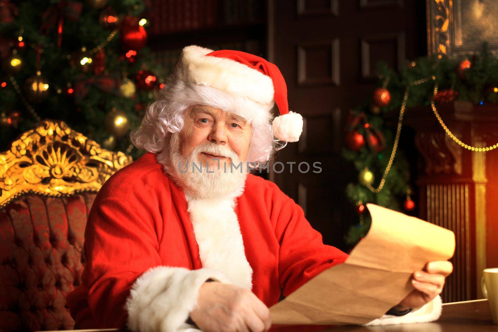 Portrait of happy Santa Claus sitting at his room at home near Christmas tree and reading Christmas letter or wish list.