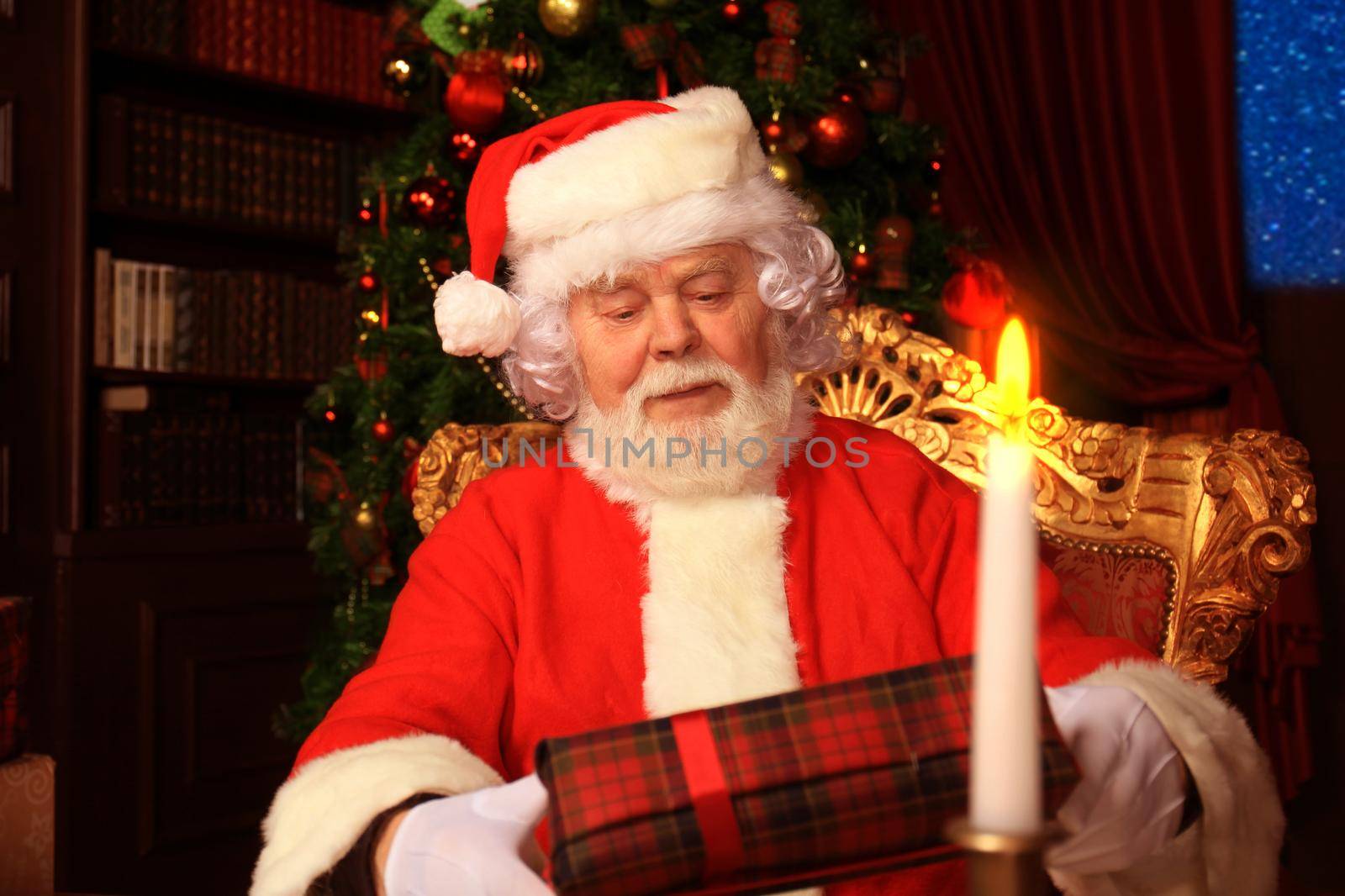 Portrait of happy Santa Claus sitting at his room at home near Christmas tree with gift box.