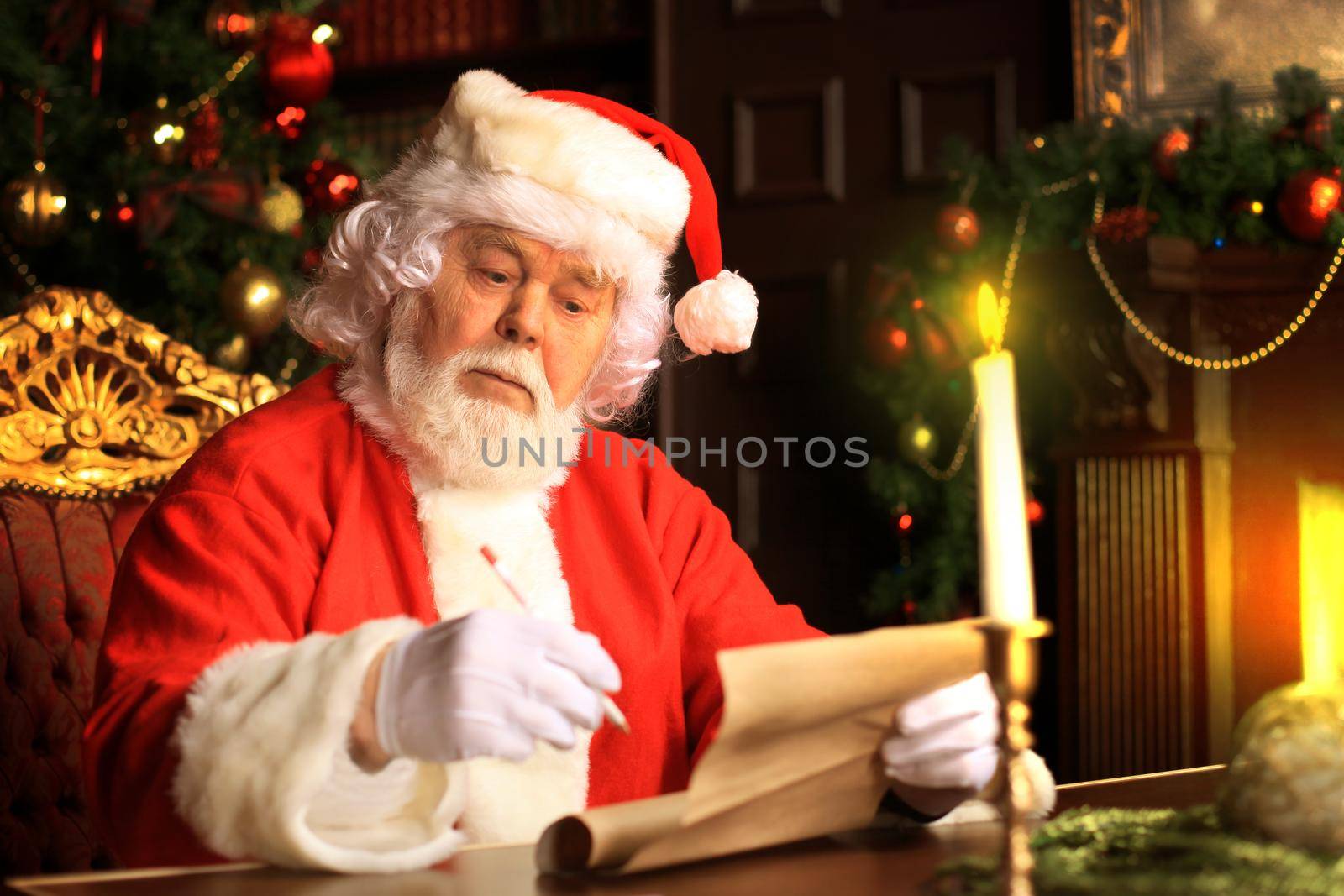 Portrait of happy Santa Claus sitting at his room at home near Christmas tree and reading Christmas letter or wish list. by tsyhun