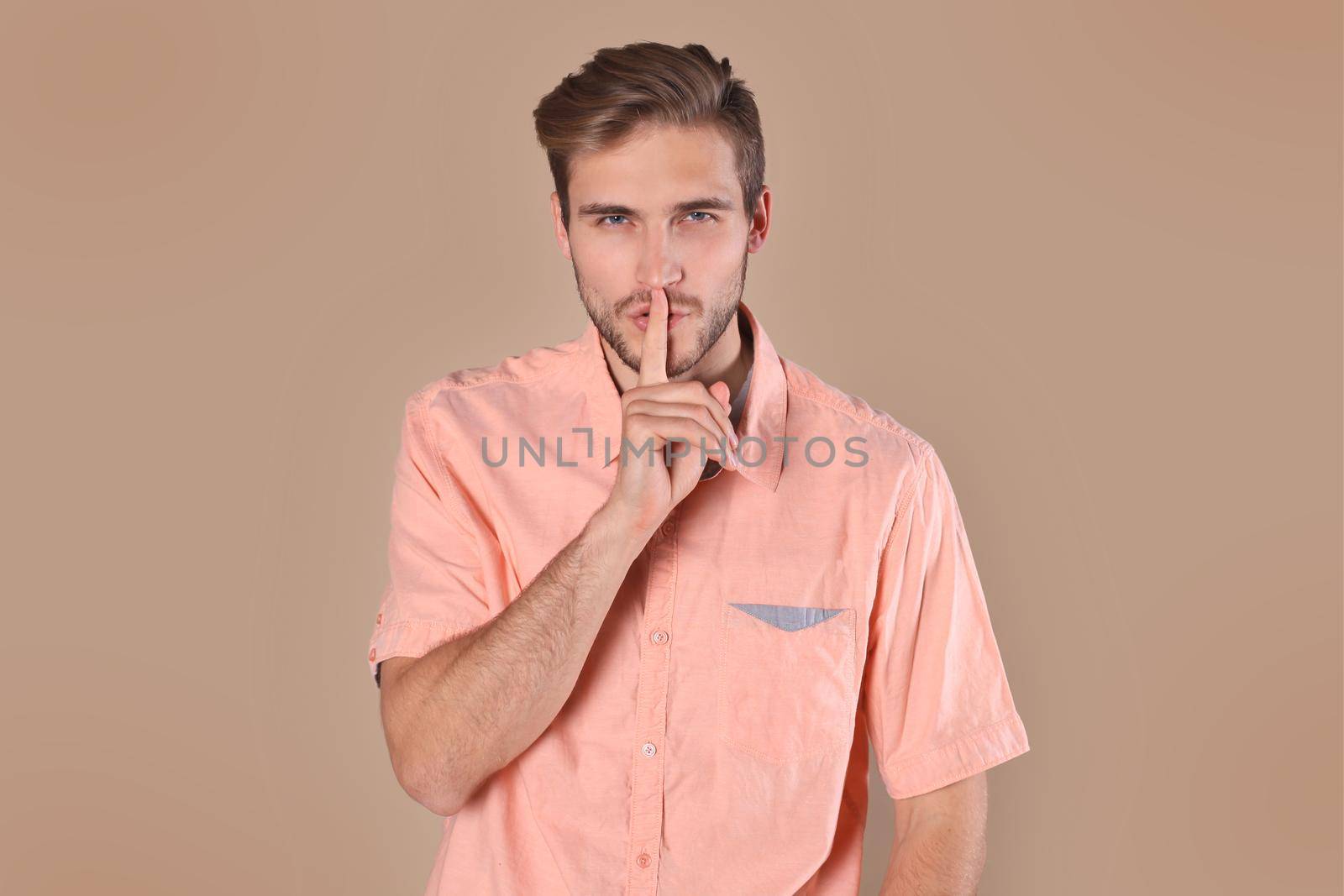 Young man with bristle posing on camera and holding index finger on lips isolated over beige background by tsyhun