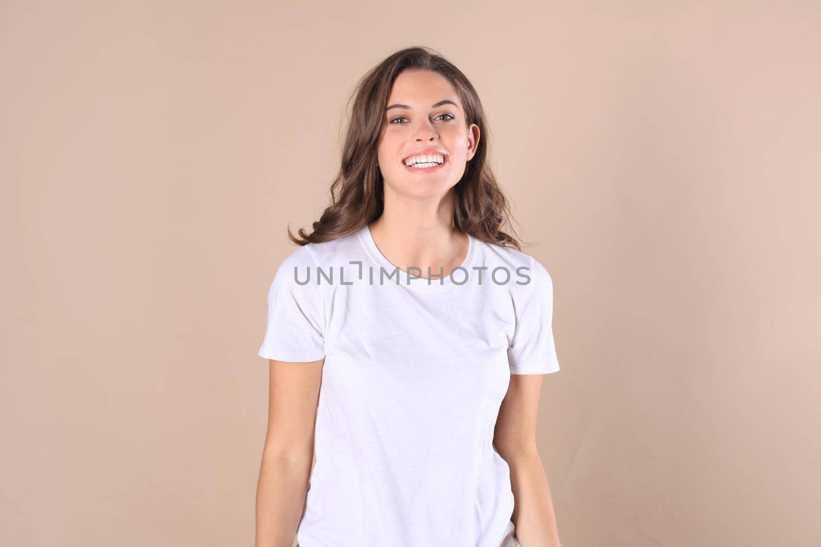 Cheerful brunette woman dressed in basic clothing looking at camera, isolated on beige background by tsyhun