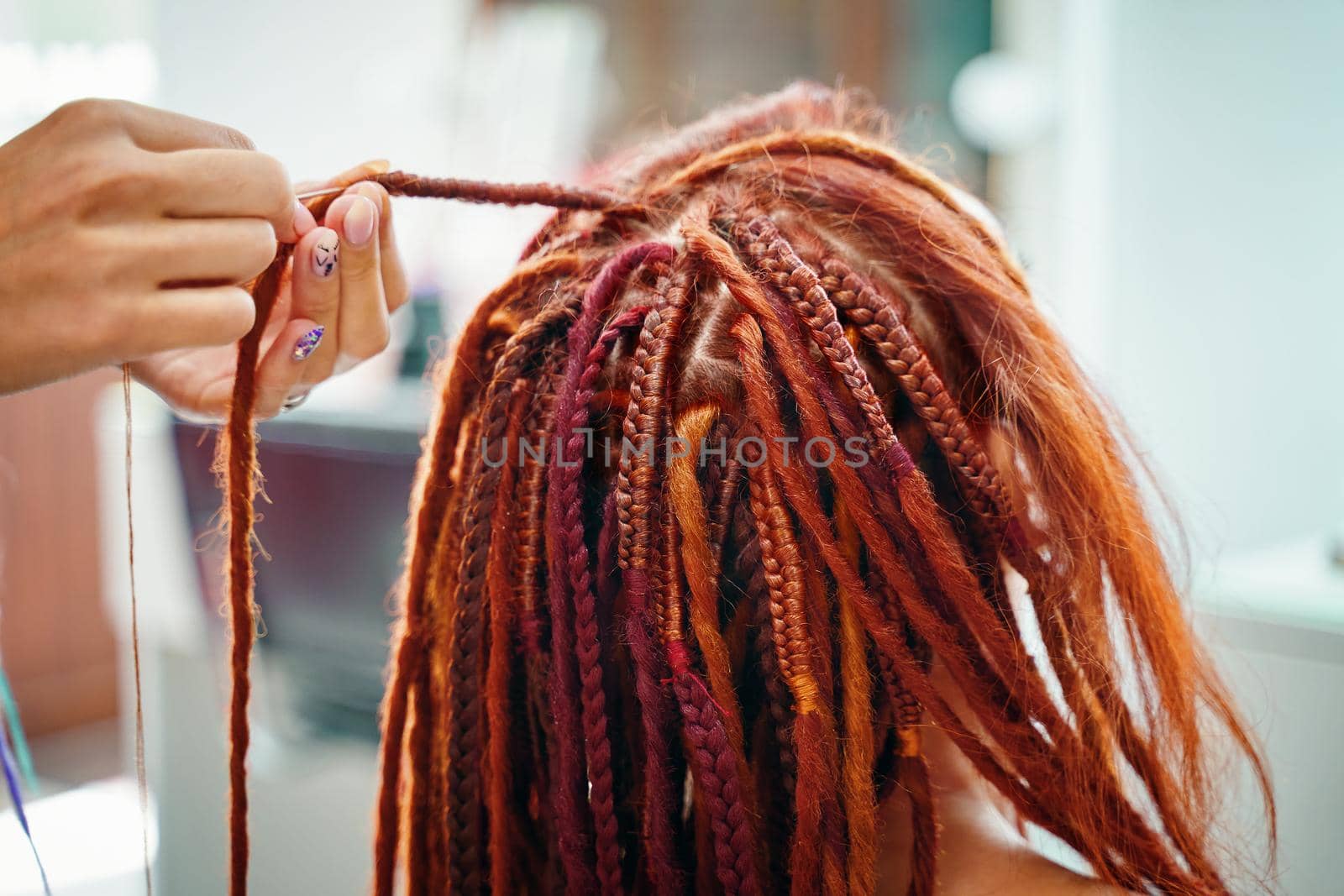 Stylish therapy professional care concept. Close up of braiding process plait with colored kanekalon. Hippie style hairstyle. Beauty salon services. Woman hairdresser weaves girl ginger dreadlocks.