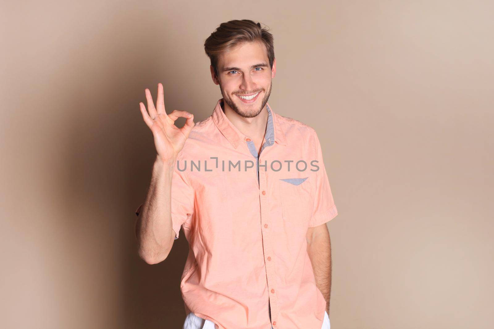 Smiling young man standing isolated over beige background, showing ok gesture by tsyhun