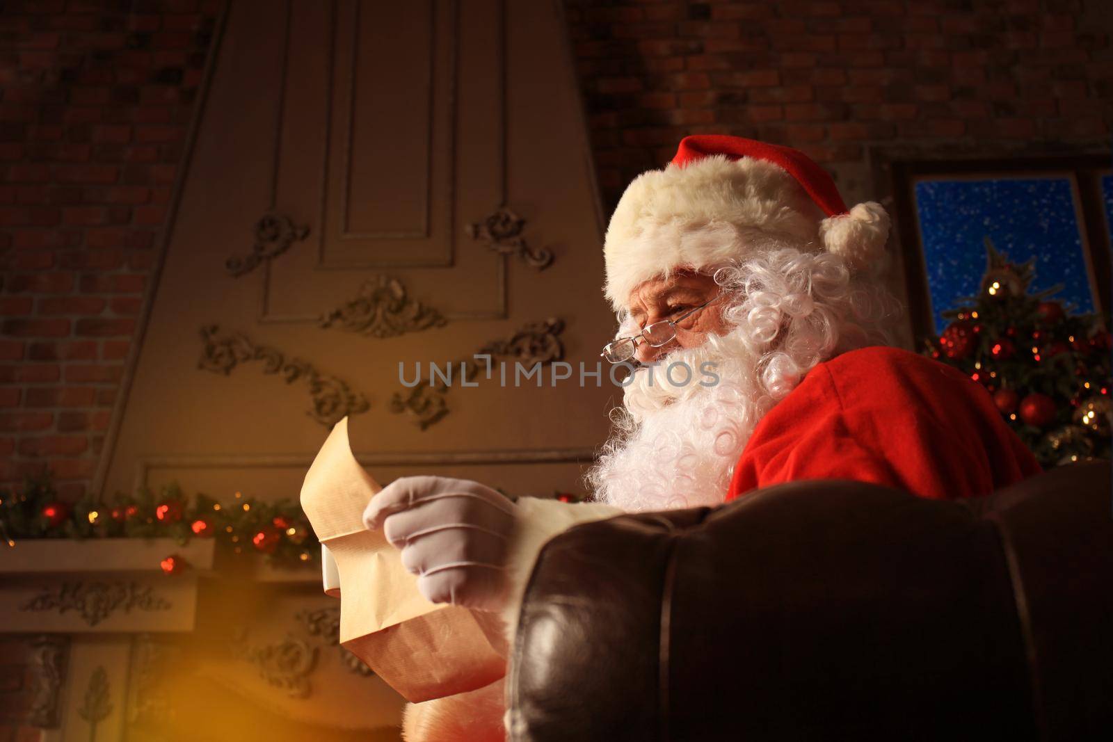 Portrait of happy Santa Claus sitting at his room at home near Christmas tree and reading Christmas letter or wish list.