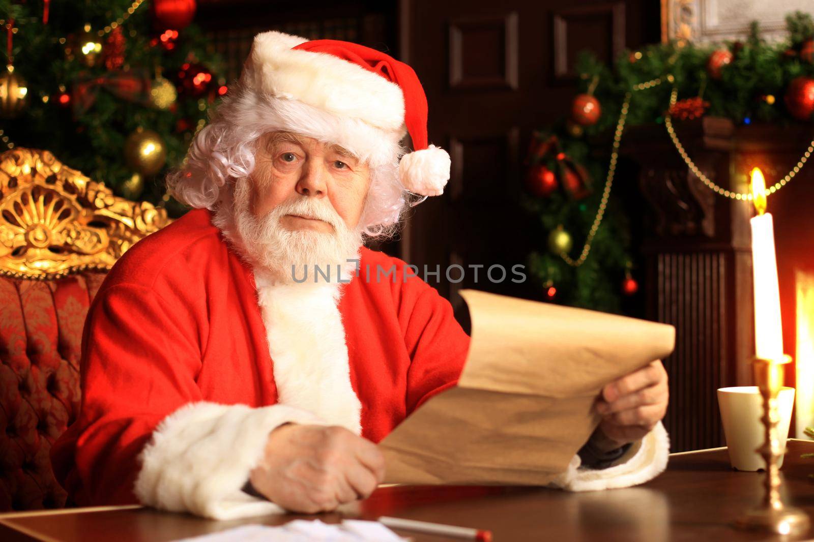 Portrait of happy Santa Claus sitting at his room at home near Christmas tree and reading Christmas letter or wish list. by tsyhun