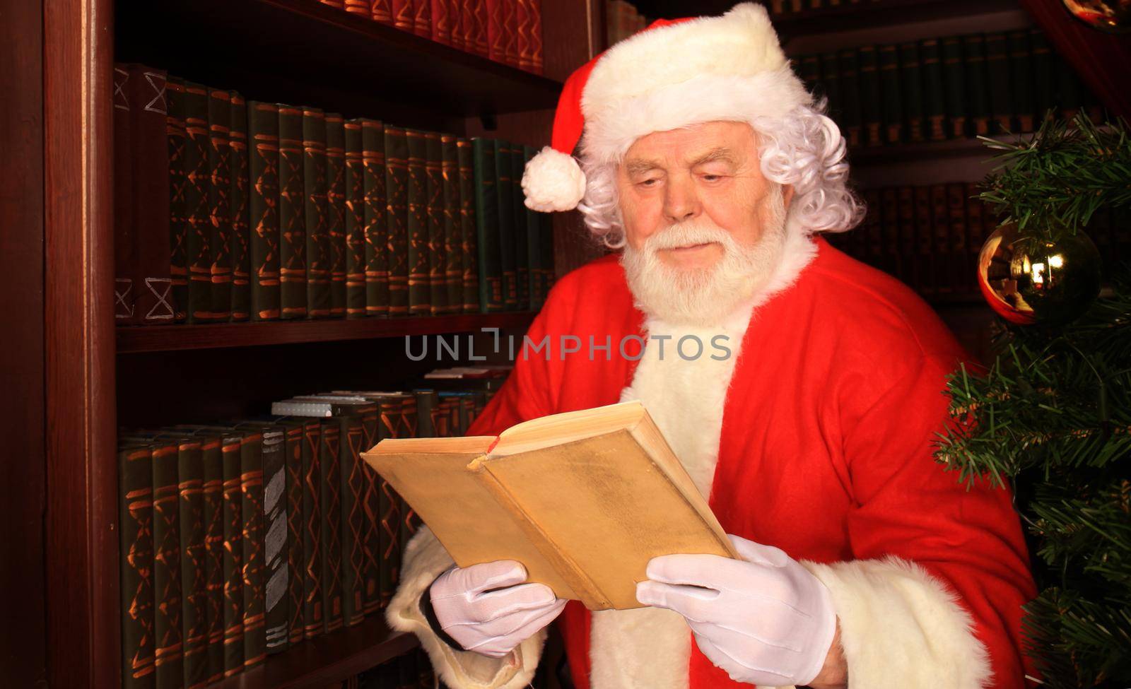 Santa standing at the Christmas tree and reading a book.
