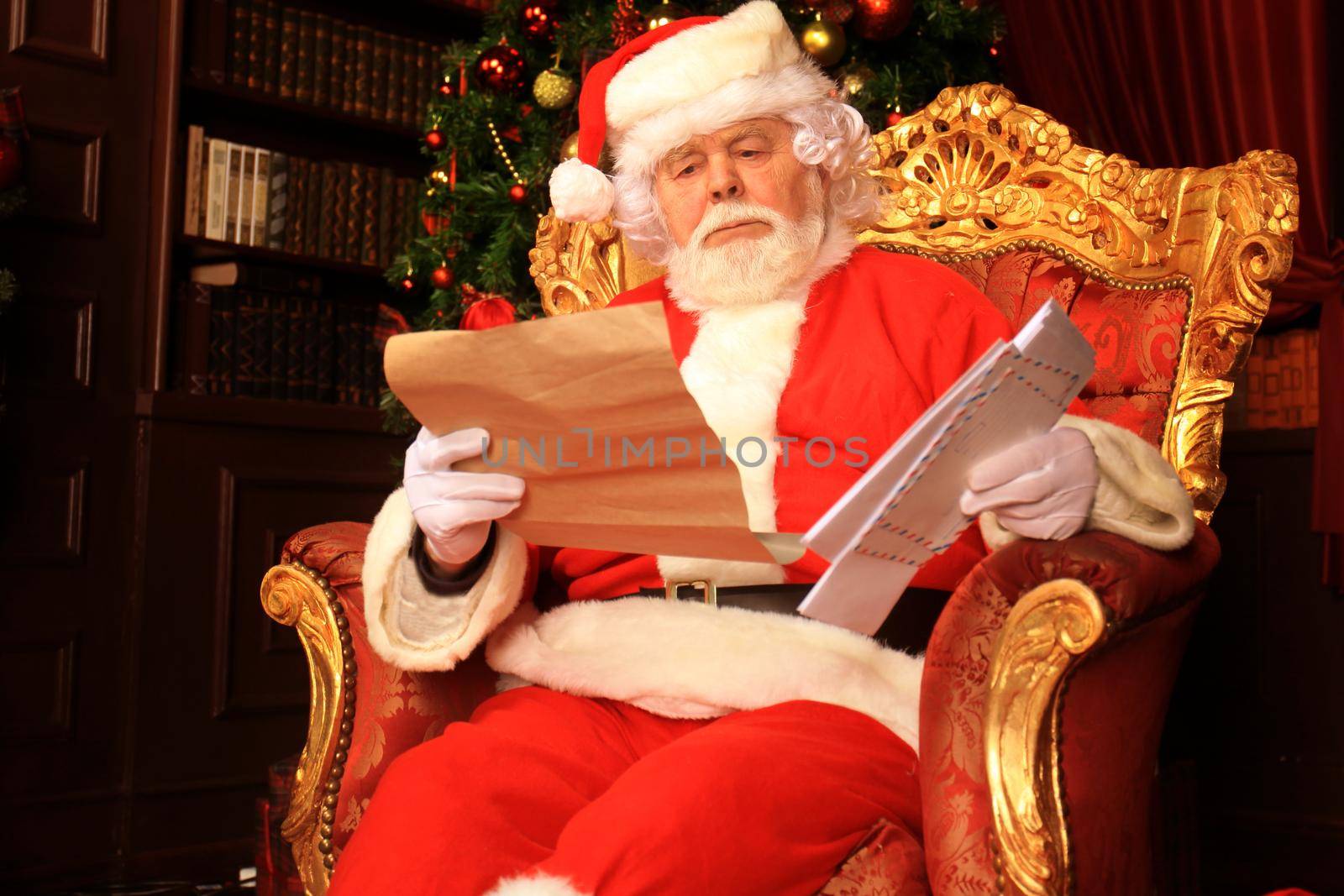 Portrait of happy Santa Claus sitting at his room at home near Christmas tree and reading Christmas letter or wish list. by tsyhun