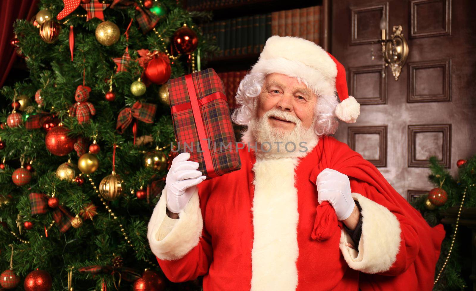 Portrait of happy Santa Claus sitting at his room at home near Christmas tree with gift box.