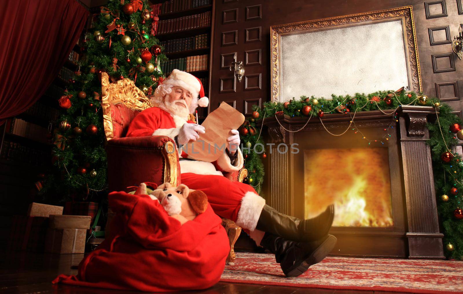 Portrait of happy Santa Claus sitting at his room at home near Christmas tree and reading Christmas letter or wish list. by tsyhun