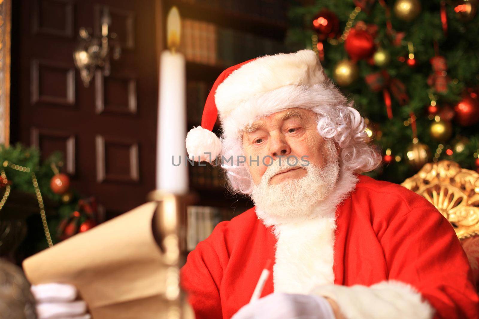 Portrait of happy Santa Claus sitting at his room at home near Christmas tree and reading Christmas letter or wish list.