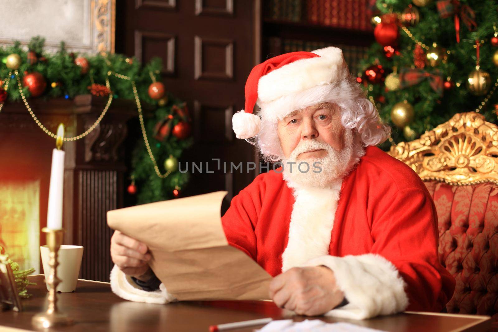 Portrait of happy Santa Claus sitting at his room at home near Christmas tree and reading Christmas letter or wish list.